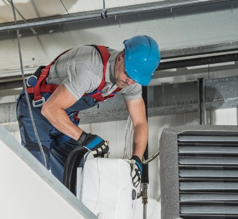 Engineer in a hard hat and safety harness maintaining a warehouse gas heater at height